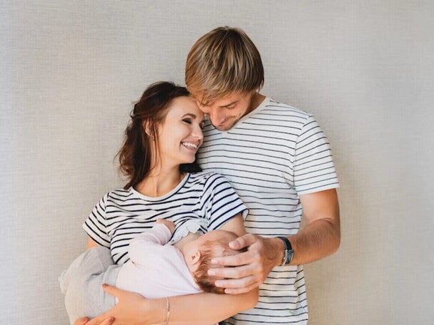 Cómo apoyar a la madre durante la lactancia Papá abrazando a mamá, quien sostiene a su bebé en brazos. Todos están sonriendo.