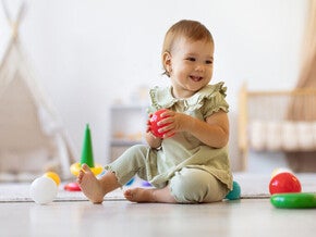 Bebé jugando con una pelota y otros juguetes en el piso, practicando habilidades motoras durante juego activo.