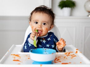 Bebé sentado en silla alta, jugando con la comida y comiendo con las manos mientras aprende a alimentarse solo durante la alimentación complementaria.