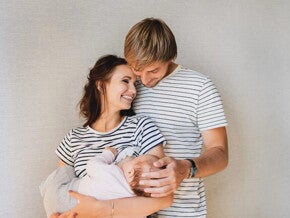 Papá abrazando a mamá, quien sostiene a su bebé en brazos. Todos están sonriendo.