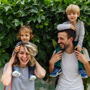 Estilos de crianza Mamá y papá al aire libre, alzando cada uno a sus hijos sobre sus hombros y sonriendo.