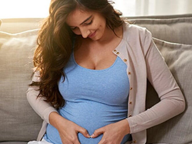Mujer embarazada acariciando su vientre, sonriendo y sentada en un sofá.