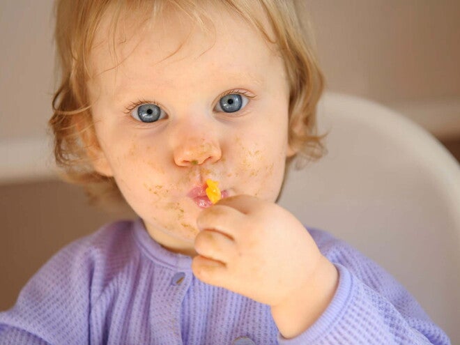 Bebé con restos de comida en la cara comiendo con una cuchara en su silla de alimentación