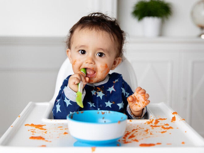 Bebé sentado en silla alta, jugando con la comida y comiendo con las manos mientras aprende a alimentarse solo durante la alimentación complementaria.
