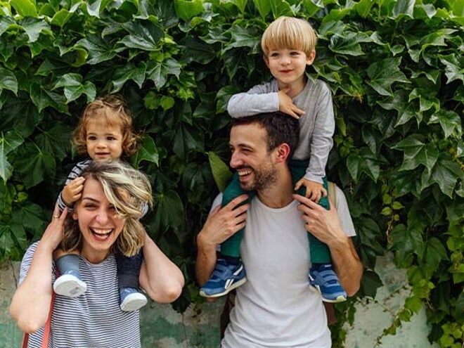 Mamá y papá al aire libre, alzando cada uno a sus hijos sobre sus hombros y sonriendo.