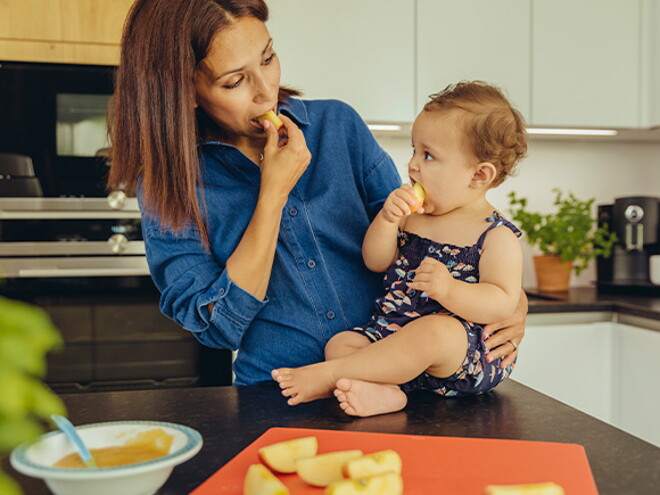Identifica cuáles son las frutas ideales para iniciar esta nueva etapa de tu bebé. Mamá y bebé comiendo fruta en la cocina.