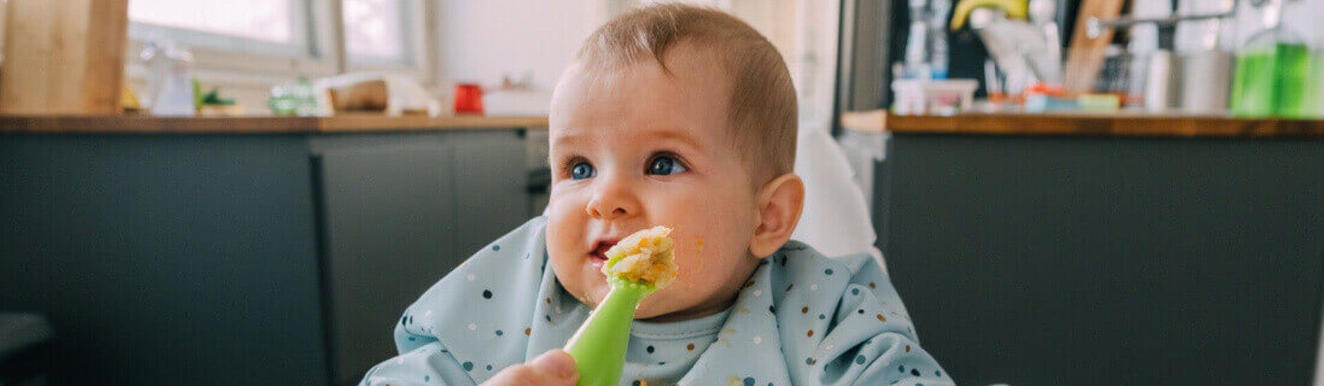 Mano de mamá dando de comer con una cuchara a su bebé que está sentado en silla alta. Alimentación complementaria del bebé.