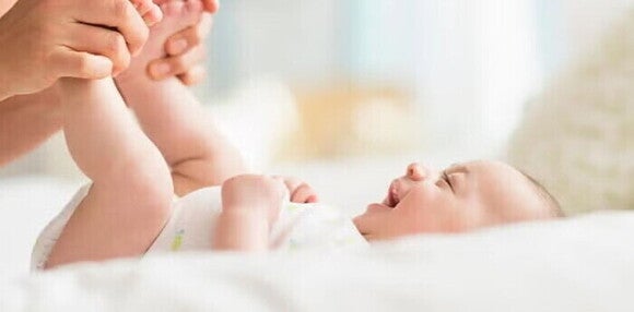 Un bebé de 2 meses desarrolla habilidades motoras finas y gruesas Bebé de dos meses riendo acostada sobre la cama mientras su mamá sostiene sus pies con sus manos.
