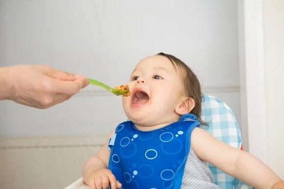 Bebé de 8 a 12 meses participando en la alimentación complementaria mientras una persona cuidadora le ofrece comida con una cuchara, promoviendo hábitos de alimentación y autonomía durante las comidas.