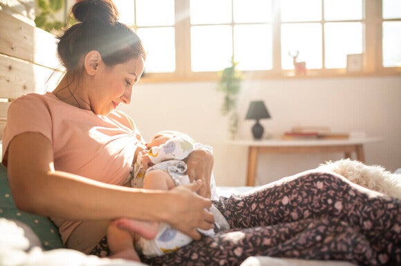 Mamá sentada en la cama, cargando a su bebé y dándole de mamar. Posición correcta para amamantar.
