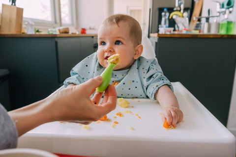 Mano de mamá dando de comer con una cuchara a su bebé que está sentado en silla alta. Alimentación complementaria del bebé.
